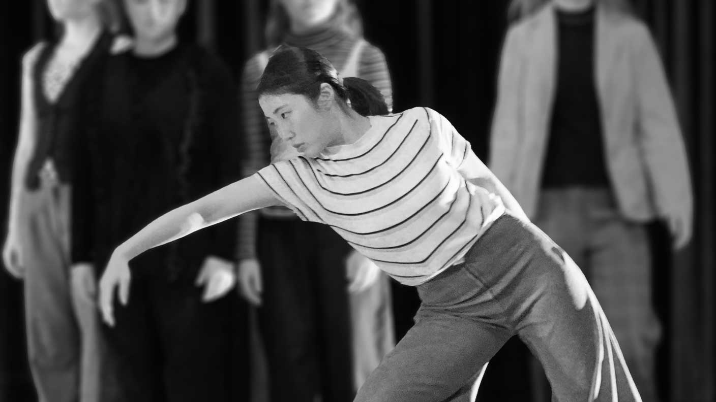 Kvinna med håret i hästsvans och vit t-shirt med svarta ränder lutar sig snett framåt med höger arm utsträckt i en danspose. I bakgrunden står fyra personer.