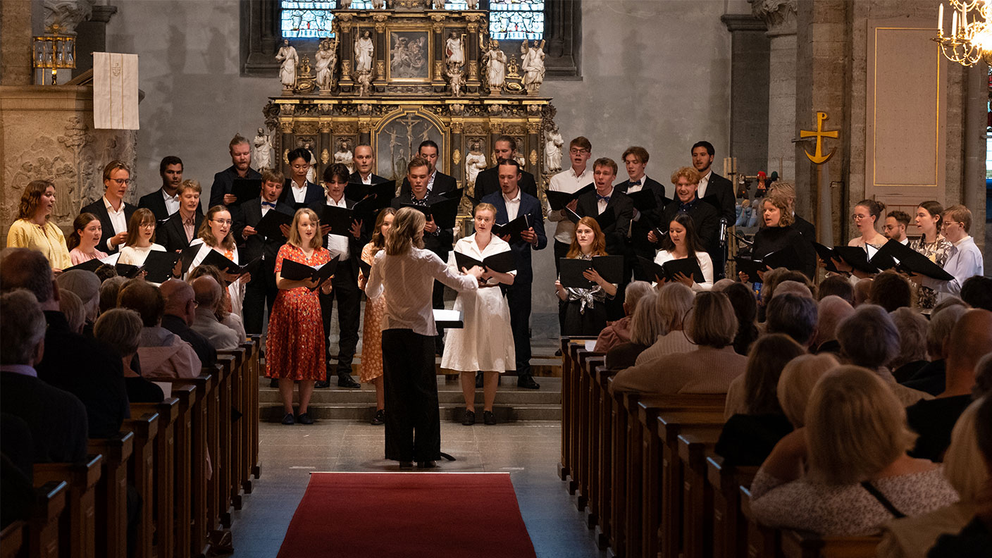 Trettiotal unga vuxna sjunger i kör ledda av kvinnlig dirigent i kyrka.