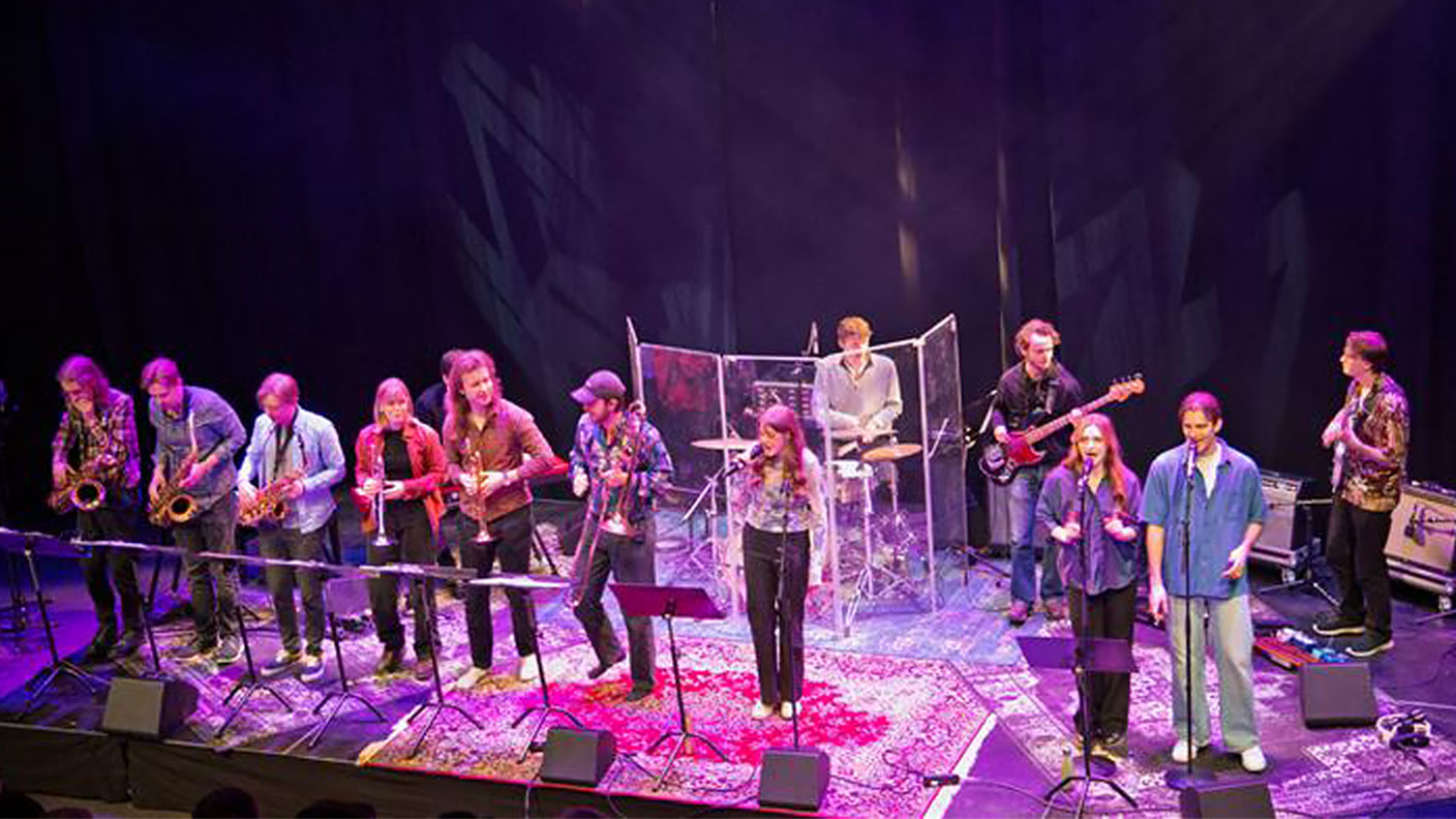 Tretton personer spelar musik på scen i lila ljussättning.