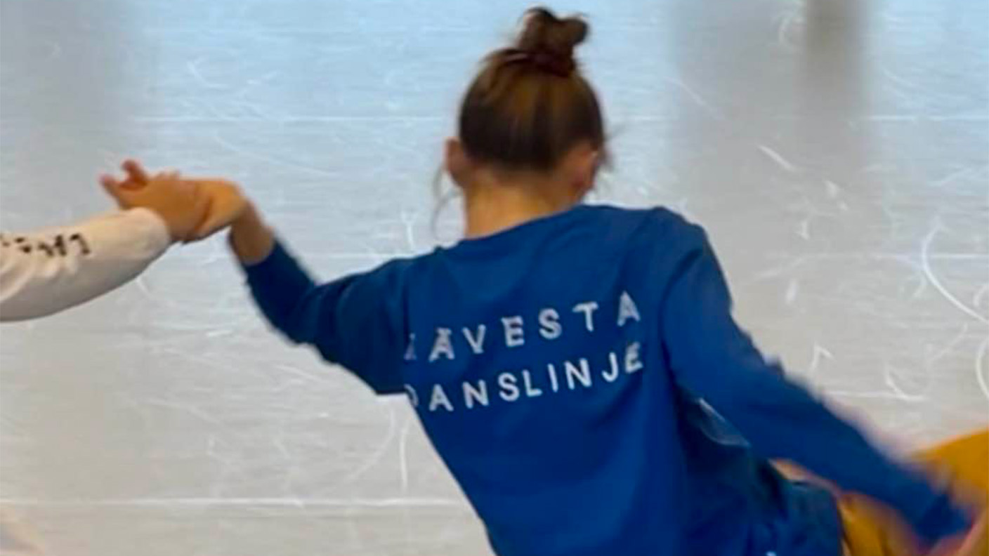 Kvinna med håret upplagt och blå collegetröja med text "Kävesta danslinje" sitter på golvet i dansrörelse.