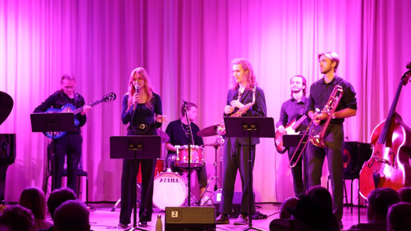 Flera personer framför musik på scen i violett ljussättning framför sittande publik.