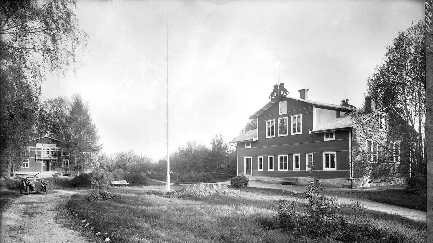 Svartvitt gammalt foto med stor träbyggnad till höger och annan byggnad längre fram till vänster.