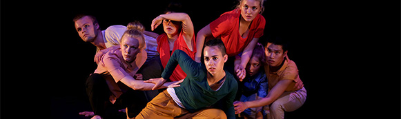 Sju danselever står i låg position i grupp och blickar åt samma håll. Svart bakgrund.