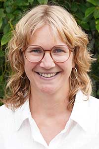 Anna har ljust axellångt hår med mittbena, hon bär bruna glasögon och en vit skjorta.