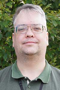 Man med gråsprängt kort hår, glasögon och grön pikétröja ler tittandes in i kameran.