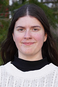 Kvinna med långt rakt mörkt hår i mittbena. Bruna ögon, näsring, svart krage under vit stickad tröja.