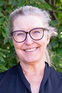 Marit har grått hår uppsatt i hästsvans. Hon har mörka glasögon och en v-ringad marinblå skjorta.