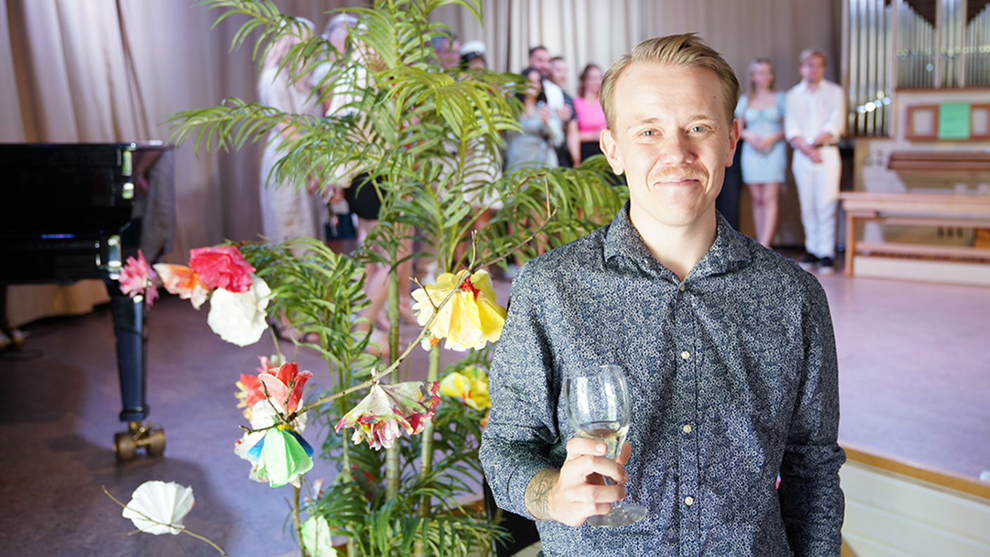 Ung man med blå blommönstrad skjorta och glasi handen ler mot kameran. I bakgrunden blommor och festklädda ungdomar på scen.