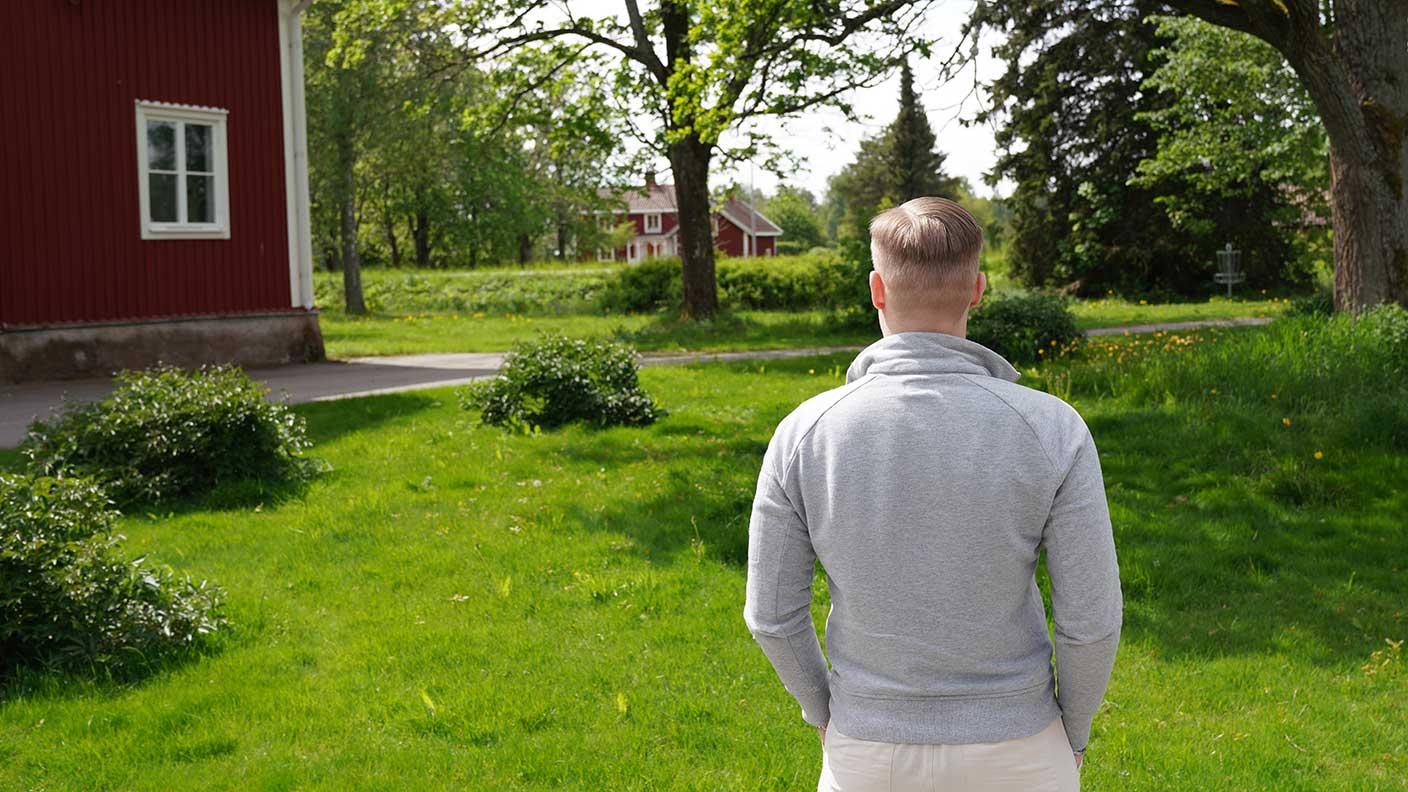 Man med kort ljust hår och grå tröja står med ryggen mot kameran och blickar ut över lantligt landskap med gröna träd och röda hus med vita knutar.
