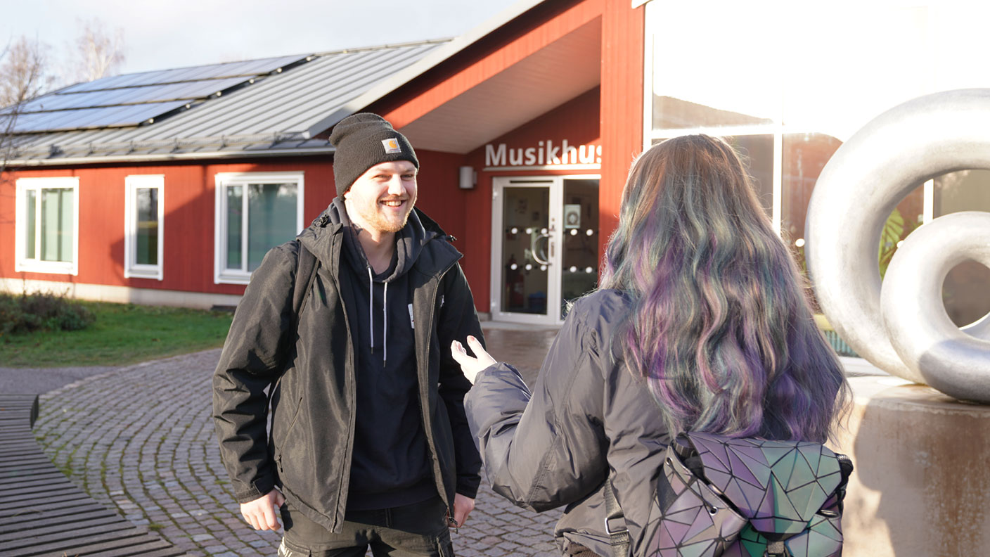 Man med mössa, svart uppknäppt jacka ovanpå blå hoodie ler och pratar med kvinna som står med ryggen till. I bakgrunden röd byggnad med texten Musikhus.