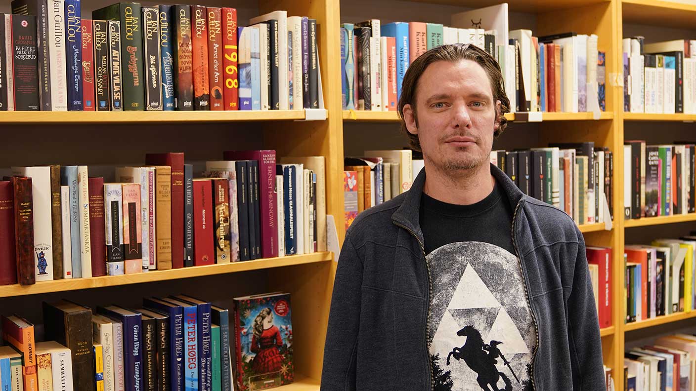 Man med mörkt bakåtkammat hår, t-shirt med tryck under mörk kofta står framför bokhylla i bibliotek.