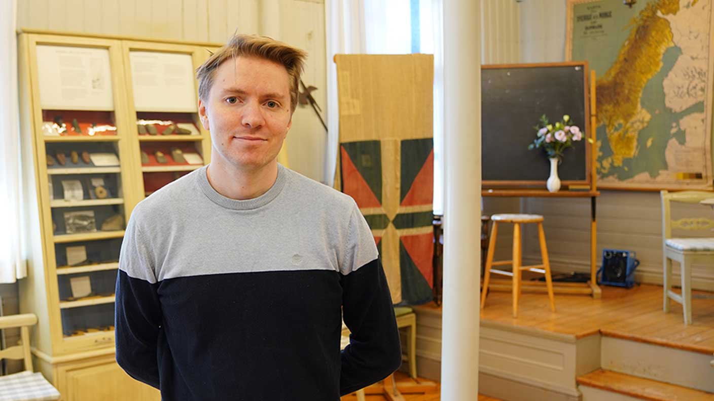 Man i kort ljusbrunt hår i halvprofil. Han har en grå och svart långärmad tröja, håller armarna bakom ryggen och ler, stående i gammaldags klassrum med fyndsamlingar och gamla kartor på väggen.