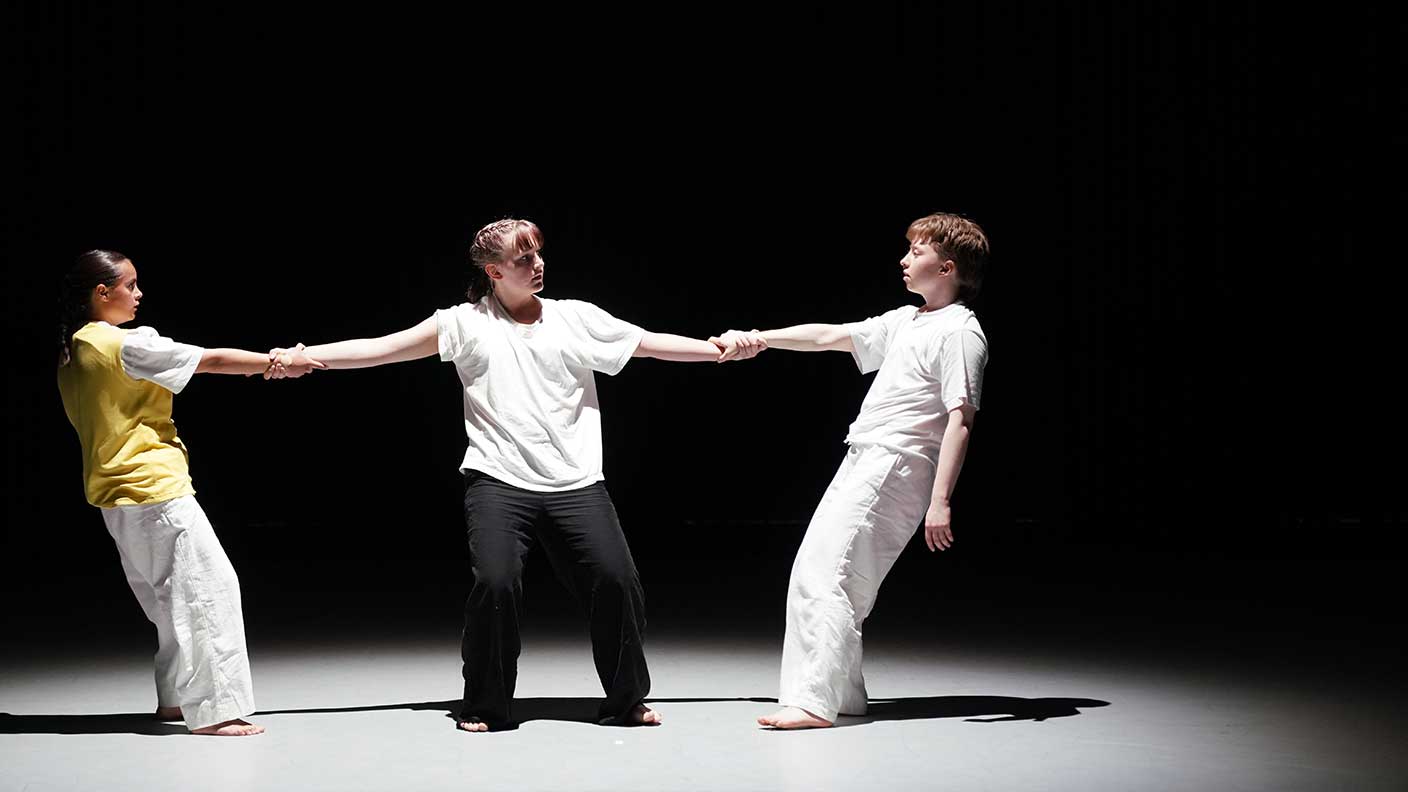 Tre personer på rad i ljuskägla i mörkt rum. Mittenpersonen håller de andra två i händerna i danspose.