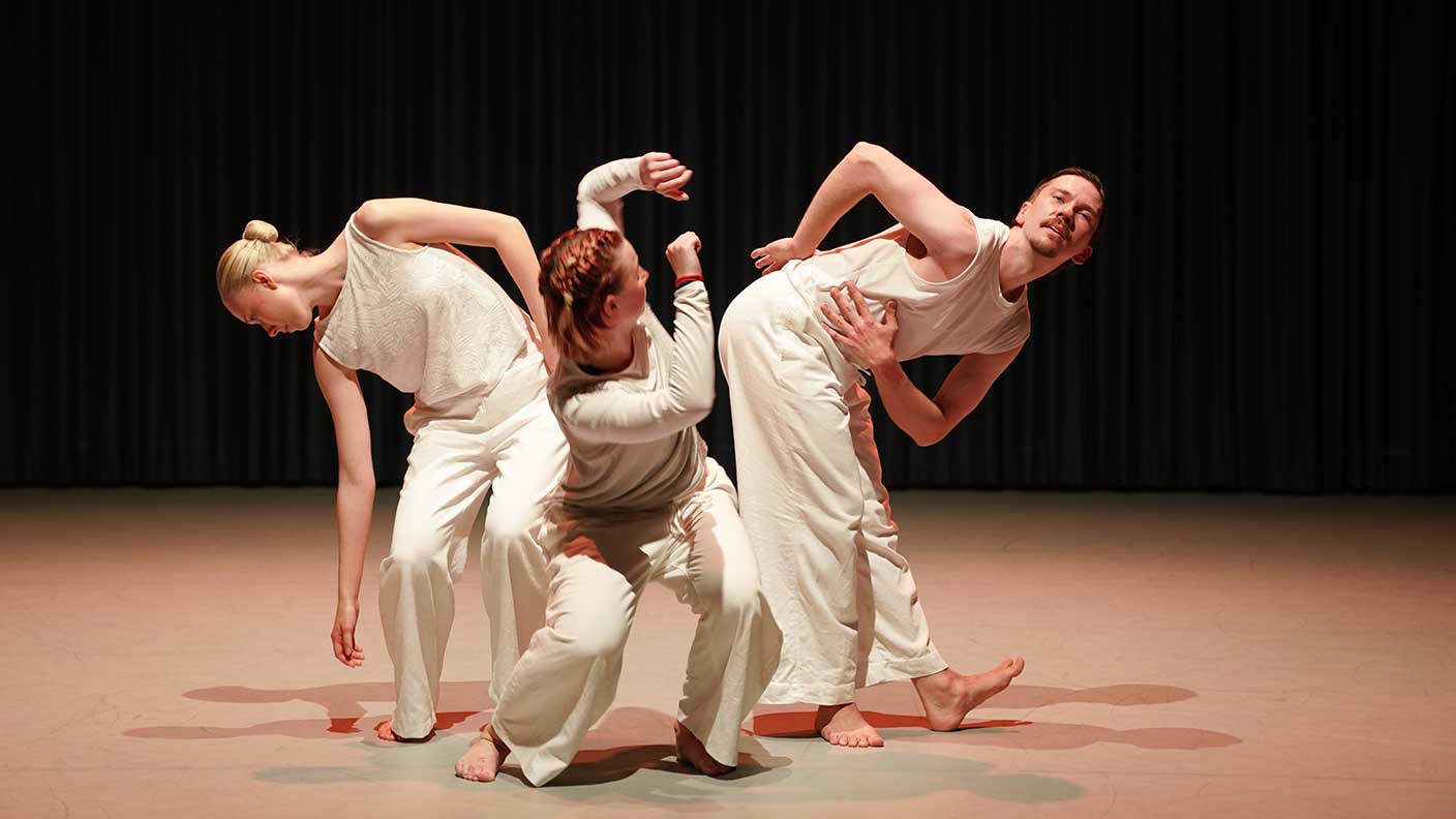Två kvinnor och en man, vitklädda stående i danspose. Mannen i sidoprofil till höger lutar kroppen framåt och har ansiktet vänt så han tittar in i bild.