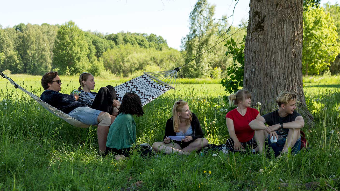 Sex personer ha samlats i en grupp ute i grönskande sommarväder. Två sitter i en hängmatta och övriga i gräset. Några tittar på något som pågår utanför bild, några talar med varandra.