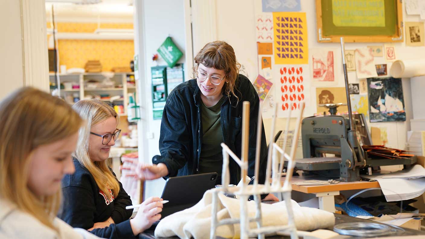 Kvinna med page, glasögon och uppknäppt mörkblå skjorta står vid bord och diskuterar med sittande kvinna som ritar med penna på skärm.