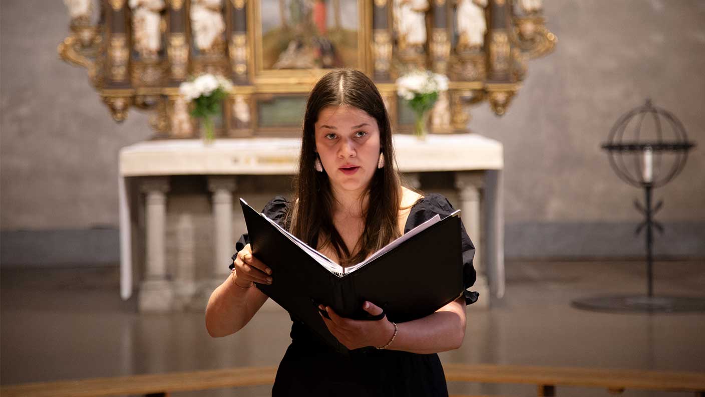 Kvinna med mörkt långt rakt hår och svart klänning sjunger hållande en svart pärm i händerna. Kyrkomiljö.