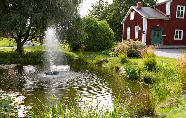 Fontän sprutar i liten damm i lummig miljö med rött trähus.