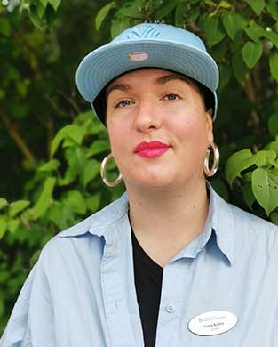 Kvinna med turkos keps, rött läppstift, stora runda örhängen och uppknäppt ljusblå skjorta över svart t-shirt.