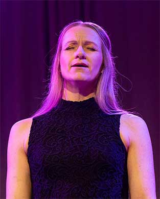 Anna-Sofia sjunger på scen. Hon har slutna ögon och halvöppen mun.  Det lila scenljuset reflekteras i hennes axellånga ljusa hår.