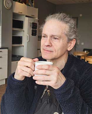 Man med grått bakåtkammat hår och mörkblå fleecejacka sitter med blicken i fjärran hållande en kopp mellan sina händer.