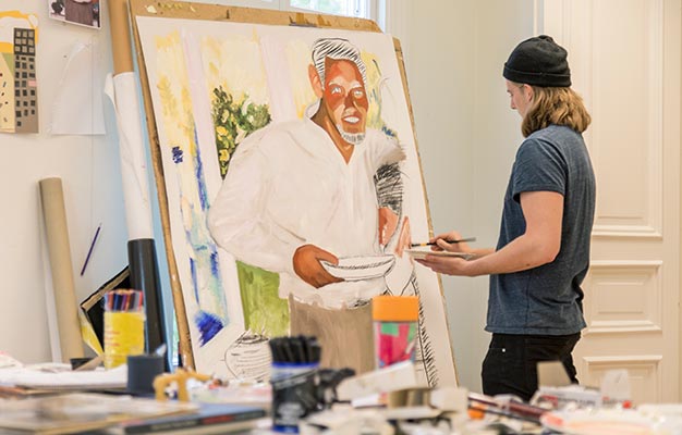 En person i gråblå t-shirt, mössa och jeans står med en pensel i handen framför en stor tavla som är halvfärdig.