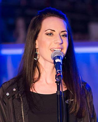 Anna står bakom en mikrofon och sjunger på scen. Hon har långt mörkt hår och bär en svart skinnjacka över en svart tröja.
