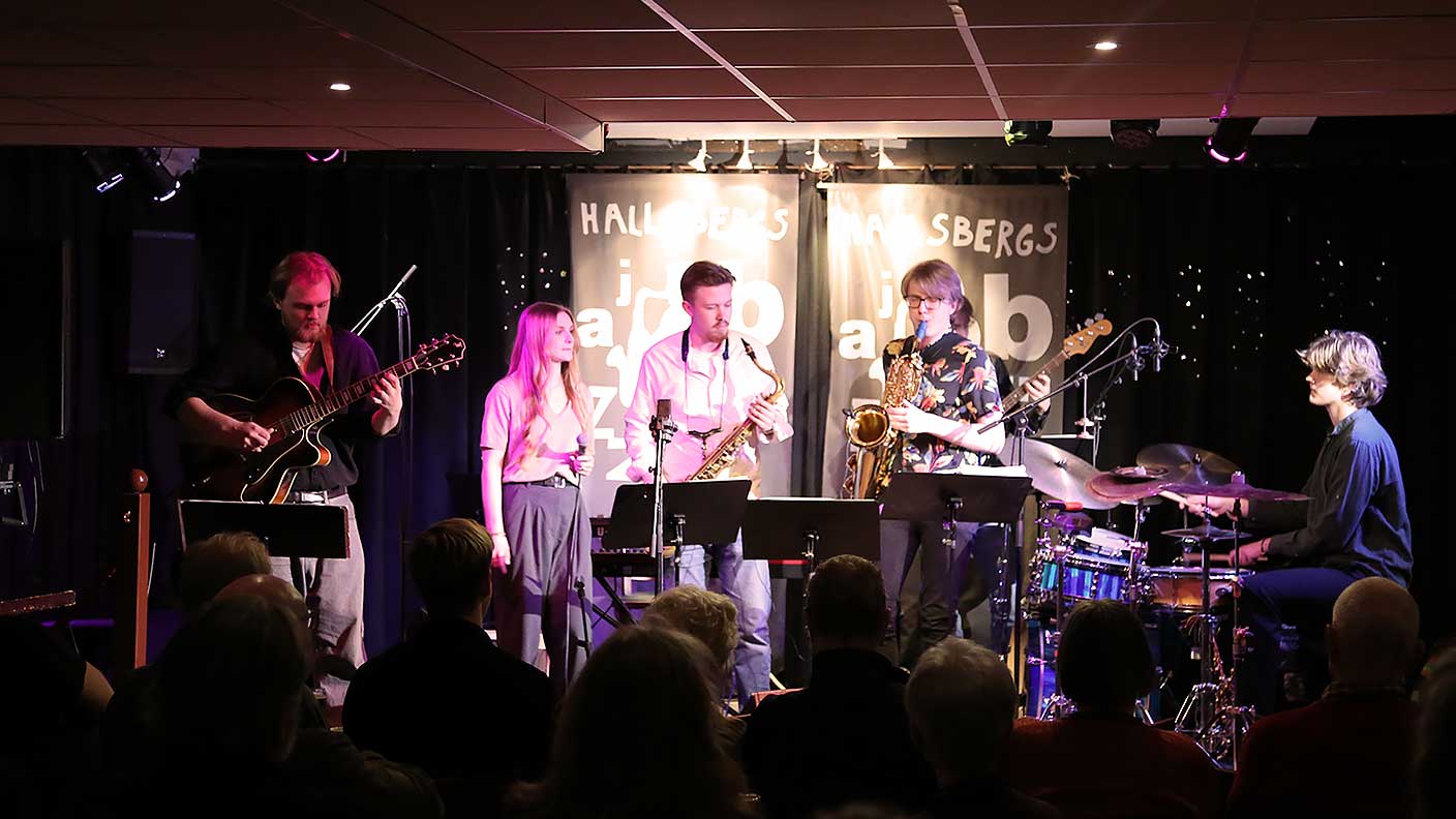 Trummis, gitarrist, basist, två saxofonister och sångare på scen framför sittande publik.