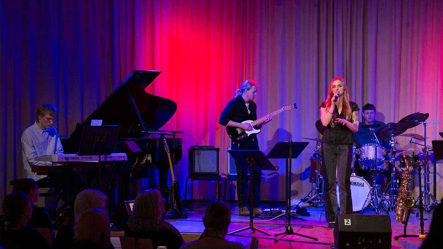 Fyra personer på scen i lilarött scenljus framför musik med sång, piano, gitarr och trummor.