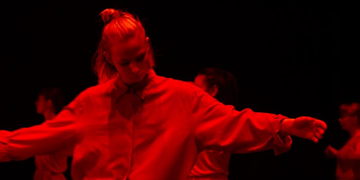 Dansare i starkt rött sken med utsträckta armar och nedböjt huvud.