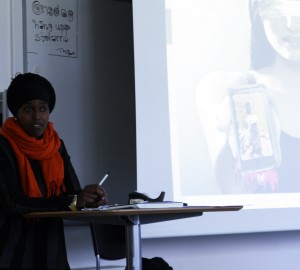 En deltagare i orange sjal och svart huvudbonad står framför en powerpoint-presentation.