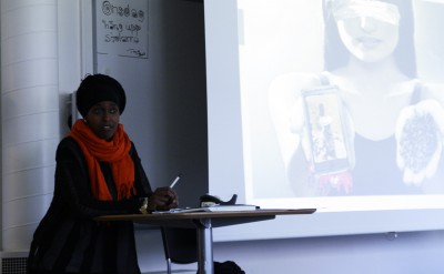 En deltagare i orange sjal och svart huvudbonad står framför en powerpoint-presentation.