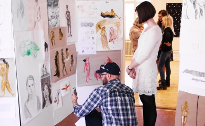 En besökare sitter på huk och fotograferar en vikvägg med kroki-teckningar. Bredvid en deltagare från konst och formgivningslinjen.