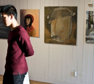 En person till vänster i bild tittar ut ur bilden, armarna bakom ryggen. I bakgrunden en vit vägg med fyra tavlor.
