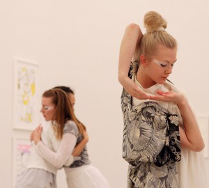 I förgrunden en dansare som fattar sina båda händer, ena armen bakom huvudet och ner mot motsatt axel. I bakgrunden två dansare i en omfamning.