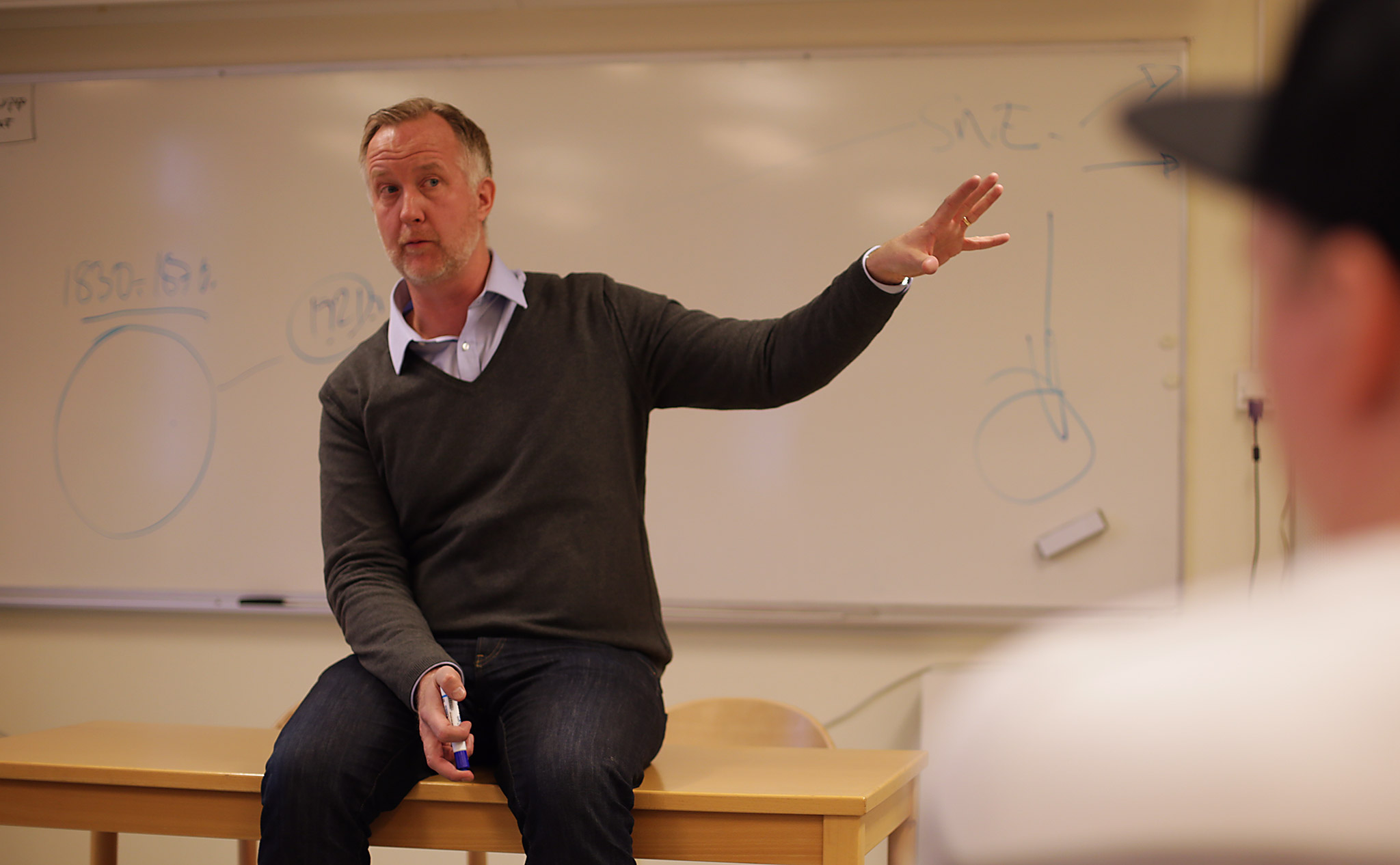 Johan Persson sitter på ett bord i en lektionssal. Pekar ut i luften.