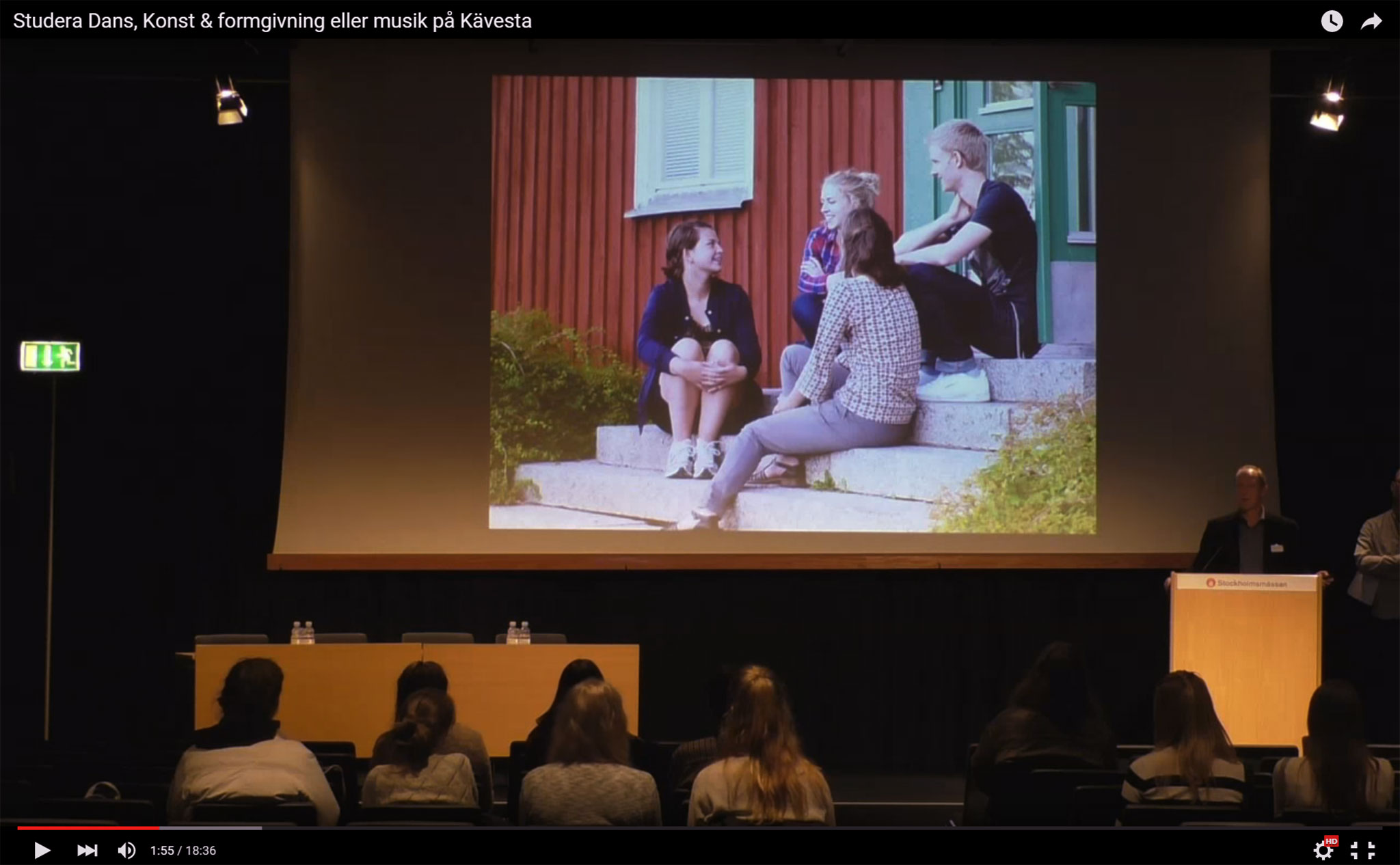 En liten publik framför en scen. På scenen en man vid en talarstol. I bakgrunden en filmduk med en bild på: tre presoner som sitter på trappen till ett rött hus.