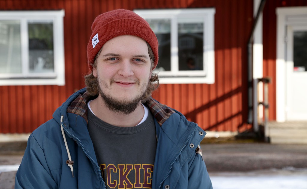 Porträtt av Oskar Eksell framför en av Kävestas röda träbyggnader. Oskar har öppen jacka och röd mössa. Snö på marken.