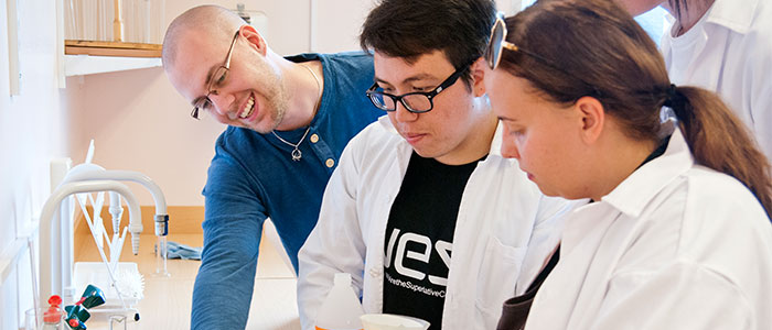 Laboration i naturvetenskapen. En lärare i blå tröja visar instruktioner för två deltagare i vita labbrockar.