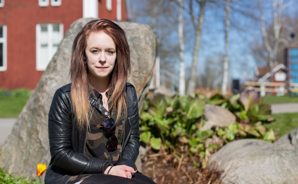 Porträttbild av Elin, som sitter framför en sten vid dammen på Kävesta folkhögskola. Elin har skinnjacka och långt hår.