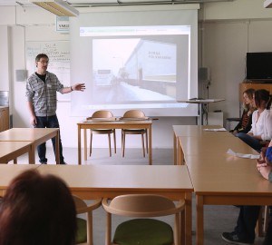 En lärare står framför en powerpointpresentation i ett klassrum. Runt ett U-format bord sitter flera deltagare och lyssnar.