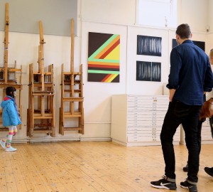 Tre personer står med ryggen mot kameran och titta på konsten som hänger på väggen: en tavla med geometriska mönster i klara färger och en tavla i svart och vitt, med abstrakt, skogsliknande motiv. Till vänster i bild går ett barn förbi med en klubba i munnen.