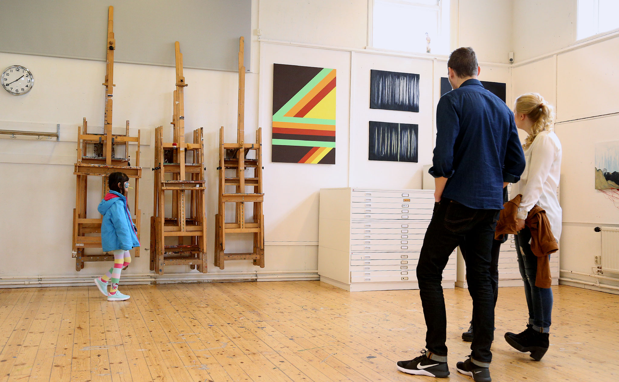 Tre personer står med ryggen mot kameran och titta på konsten som hänger på väggen: en tavla med geometriska mönster i klara färger och en tavla i svart och vitt, med abstrakt, skogsliknande motiv. Till vänster i bild går ett barn förbi med en klubba i munnen.