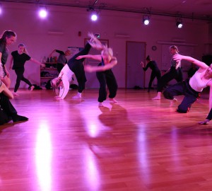 Dansare utspridda på scen i lila ljus. Många är suddiga av rörelse.
