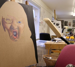 Till vänster en stor påbörjad tavla med en person som gör en grimas. I bakgrunden arbetsbänkar fyllda av olika prylar och projekt.