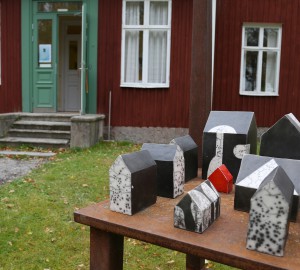 Flera små hus i keramik står på en överdimensionerad stol i metall.