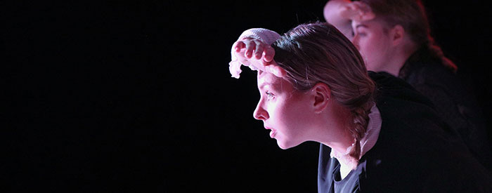 Foto från en dansföreställning. Kristin i profil spejar åt vänster i bild, med ena handen skuggar hon ögonen.