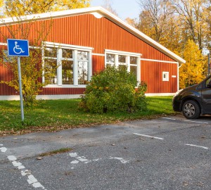 Låg vy över en parkeringsruta och en skylt med symbolen för handikapparkering. I bakgrunden ett lågt rött hus - danshuset.