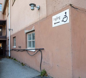 Husgavel med skylt med handikappsymbol. Bakom den leder en ramp ner till en dörr.