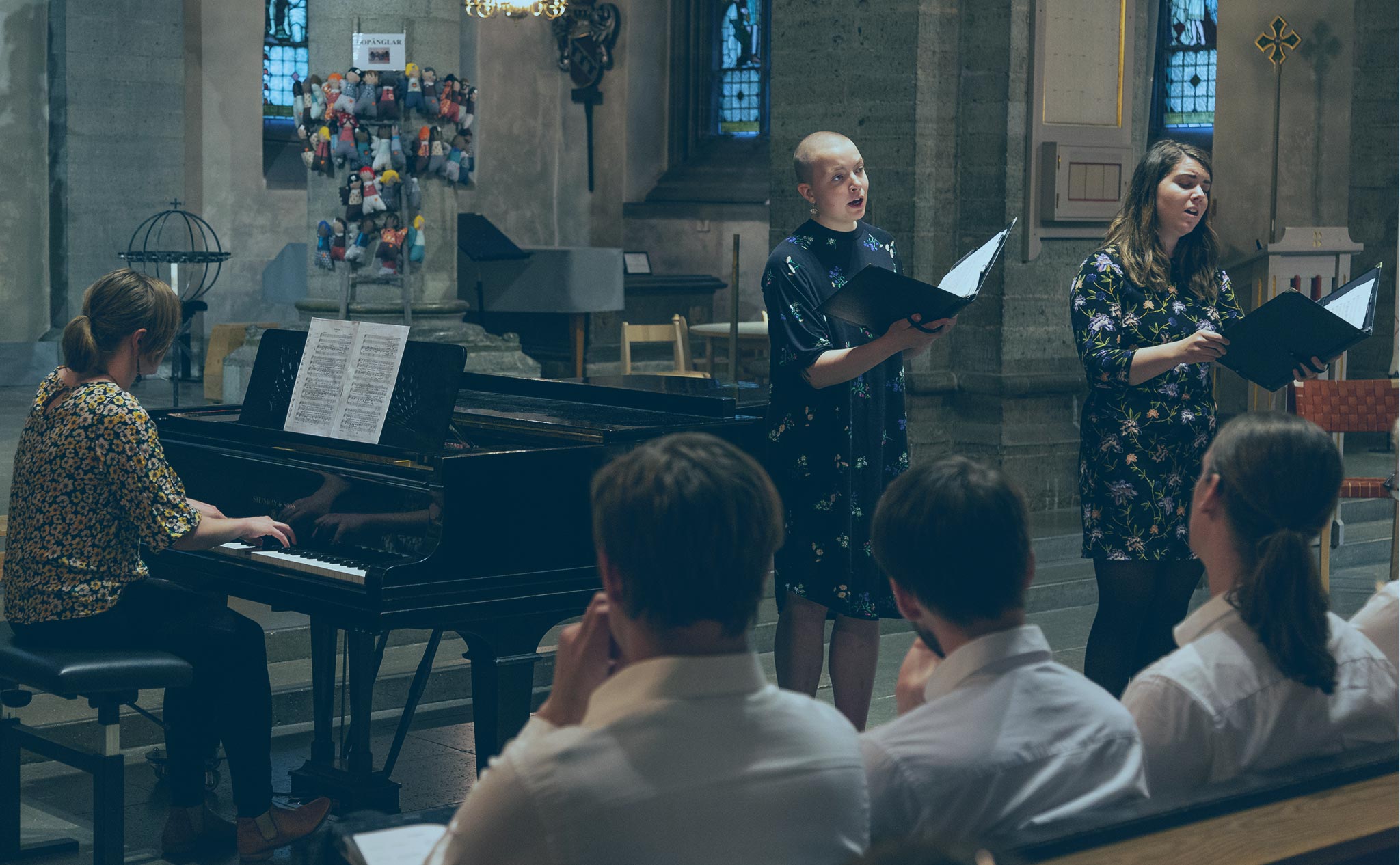 Blåtonat foto från Nicolaikyrkan i Örebro. En pianist ackompanjerar två sångare. I förgrunden tre personer i publiken klädda i vita skjortor.
